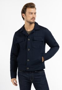 Dunkelblaues Hemdjacke aus strukturiertem Stoff, mit zwei Brusttaschen, einem Reverskragen und einem geraden Saum. Getragen mit dunklen Jeans.