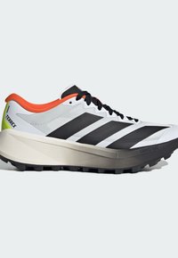 Chaussures de course avec un schéma de couleurs blanc, noir et orange. Elles comportent une semelle intermédiaire épaisse, une tige texturée et une semelle extérieure en caoutchouc noir avec des crampons pour la traction.
