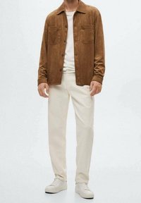 Brun ruskindsjakke med to brystlommer, hvid t-shirt med rund hals nedenunder, lyscremefarvede bukser og lyse sneakers.