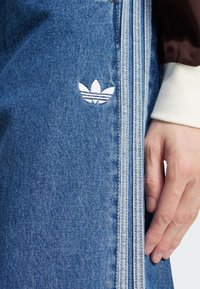 Close-up de jeans de ganga azul com o logótipo da Adidas e três riscas azul claro, ao lado de uma mão que usa uma manga castanha com uma bainha branca.