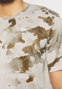 Szara koszulka typu T-shirt w kamuflażu z jasnymi i ciemnymi brązowymi wzorami, krótkimi rękawami, okrągłym dekoltem oraz małym białym logo Nike na piersi.