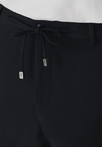 Czarne spodnie z elastycznym pasem, z niebieskim guzikiem, metalowymi akcentami na końcach sznurka oraz gładkim, teksturowanym materiałem.