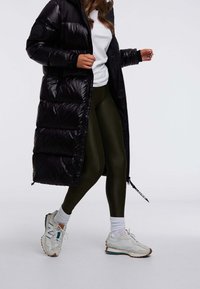 Zwarte pufferjas met glanzende afwerking, met ritssluiting, groene leggings en witte sneakers met gekleurde accenten, gezien in een lopende pose.