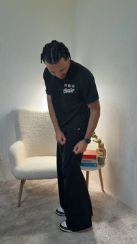 Άνδρας με πλεγμένα μαλλιά ρυθμίζει ένα φαρδύ μαύρο παντελόνι, στεκόμενος κοντά σε μια λευκή υφασμάτινη πολυθρόνα και ένα μικρό στρογγυλό τραπέζι με βιβλία και ένα βάζο.