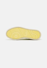 Suola di una sneaker con fondo in gomma gialla testurizzata, caratterizzata da motivi circolari e lineari, contornata da un bordo beige chiaro.