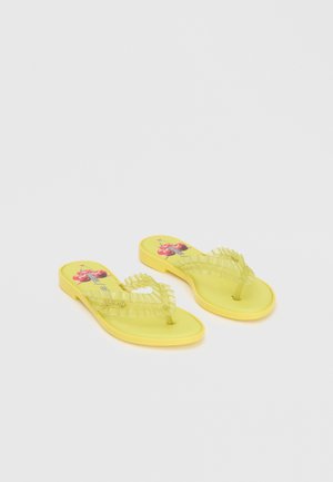 Sandales plates jaunes avec des brides transparentes froncées et des motifs cerises sur la semelle, positionnées côte à côte sur un fond uni.