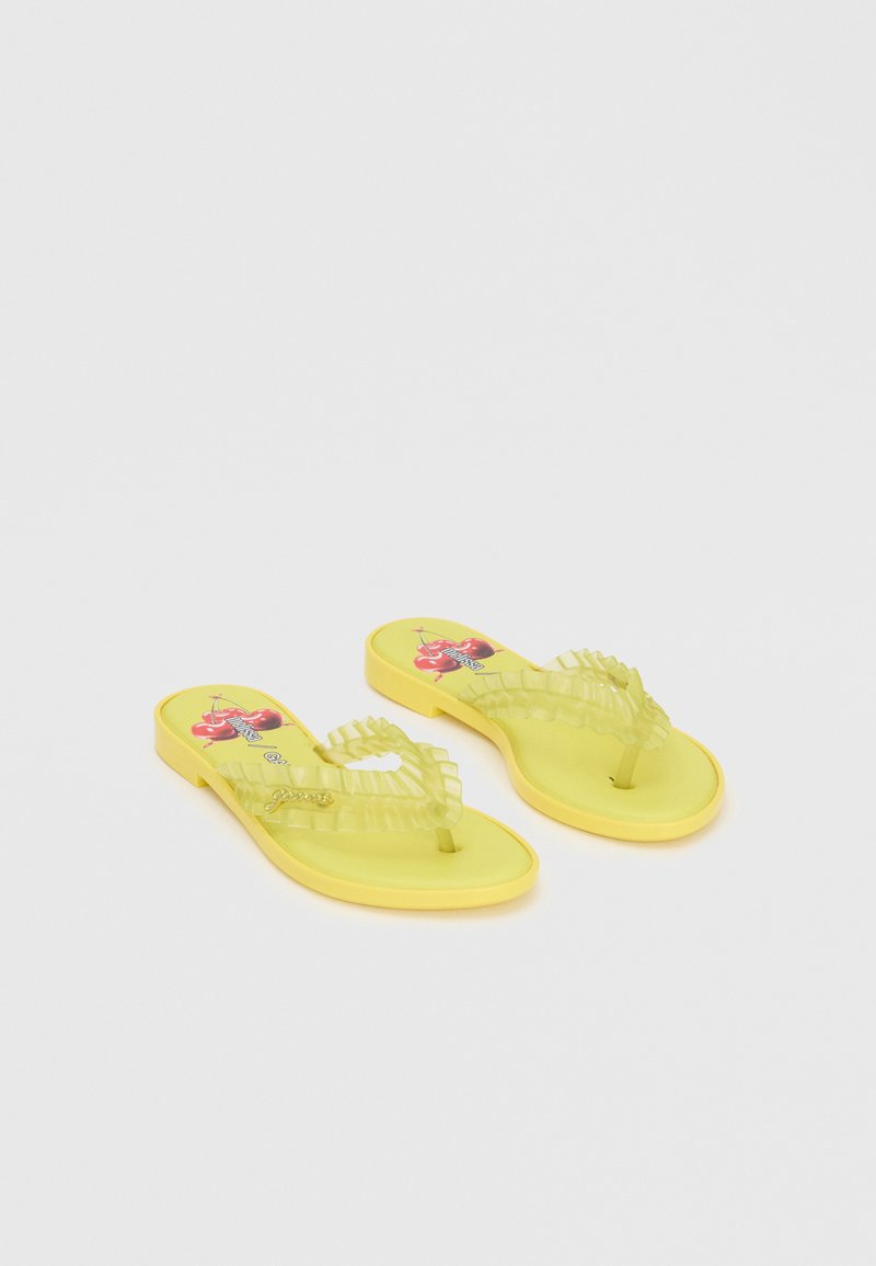 Sandales plates jaunes avec des brides transparentes froncées et des motifs cerises sur la semelle, positionnées côte à côte sur un fond uni.
