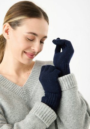 Marineblaue Strickhandschuhe mit gerippten Bündchen werden an den Händen getragen, kombiniert mit einem hellgrauen V-Ausschnitt-Pullover mit strukturiertem Muster.