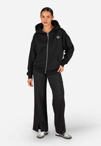 Čierna mikina na zips s predným vreckom, spárovaná s čiernymi voľnými nohavicami. Materiál vyzerá mäkko a pohodlne, s minimálnym zdobením.