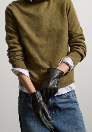 Guantes de cuero negro con acentos de botones, usados sobre un suéter verde y una camisa blanca, presentan un diseño ceñido y una textura suave.