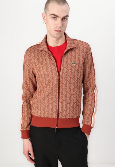 Veste zippée marron avec un motif géométrique, poignets et ourlet côtelés, accents rouges, et un petit logo vert sur la poitrine. Pantalons noirs.