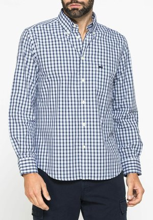 Uomo che indossa una camicia a quadri blu e bianca con bottoni e maniche lunghe e pantaloni scuri, in piedi di fronte a uno sfondo bianco.