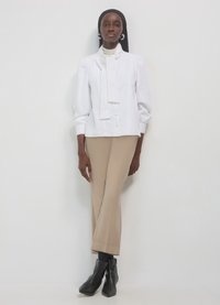 Weiße Bluse mit Schleife, gerückten Ärmeln und Knopfleiste; kombiniert mit beigen, weit geschnittenen Hosen und schwarzen Stiefeletten.