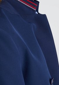 Navy blue jacket with a structured collar featuring red and white stripe accents. Smooth fabric with a button detail on the front.