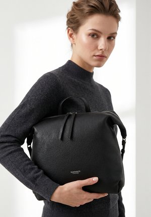 Jonge vrouw in een donkere trui houdt een grote zwarte leren Diana&Co Firenze handtas met dubbele ritssluitingen vast.