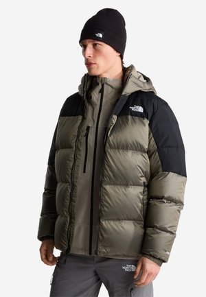 Uomo con berretto nero, giacca imbottita North Face verde oliva e nera, e pantaloni grigi North Face, in piedi davanti a uno sfondo uniforme.