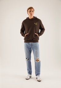 Felpa con cappuccio marrone con tasca frontale e logo giallo, abbinata a jeans chiari strappati e scarpe da ginnastica bianche e nere.