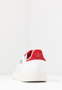 White Adidas sneaker with red heel tab, visible Adidas logo, rubber sole, and textured side panels, angled to show rear and side details.