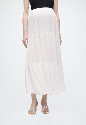 Naine kannab voolavat valget kihilist midi seelikut, musta varrukateta toppi ja beežikaid rihmikuga kontsaga sandaale, taust on valge ja ühtlane.