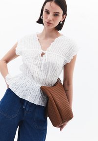 Blusa bianca plissettata con maniche corte arricciate, dotata di chiusura a fiocco. Abbinata a jeans blu a vita alta e una borsa intrecciata marrone.