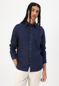Camicia a manica lunga blu navy realizzata in tessuto leggero, con colletto a camicia e taschino sul petto, indossata con pantaloni chiari.
