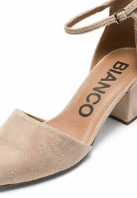 Zapato de ante beige con tacón cuadrado y diseño de punta. Presenta una correa al tobillo con un hebilla metálica. Logo impreso en la plantilla.