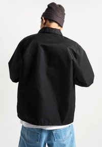 Black jacket with a relaxed fit, lightweight material, and a collar. Paired with light blue jeans, viewed from the back. Ribbed beanie on head.