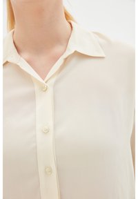 Femme portant un chemisier beige à boutons avec un col pointu, gros plan sur la partie supérieure de la poitrine et le cou.