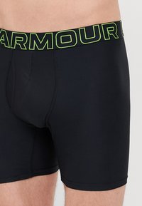 Schwarze Boxershorts aus glattem Stoff mit einem breiten Bündchen, auf dem in grüner Schrift "ARMOUR" steht. Das Design ist mit einem konturierten Pouch ausgestattet.