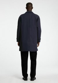 Mornarska jakna ravnega kroja z širokim ovratnikom, gumbi na manšetah in gladko teksturo, oblečena čez črne hlače in čevlje.