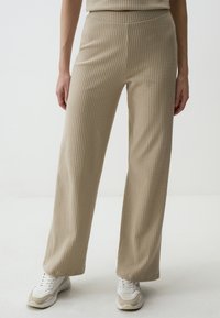 Beige ribbed knit pants with a high waist, wide legs, and a textured finish, paired with white and beige athletic shoes.