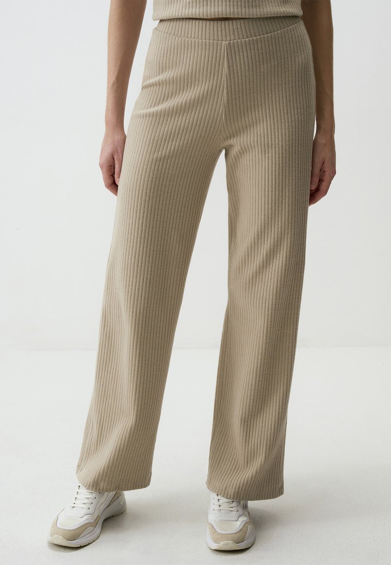 Beige ribbed knit pants with a high waist, wide legs, and a textured finish, paired with white and beige athletic shoes.