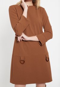 Brun figurnära klänning med trekvartslånga ärmar, rund halsringning, två framfickor med bärnstensfärgade ringar och strukturerad tyg.