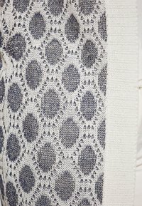 Tessuto a maglia bianco con un motivo testurizzato che presenta forme geometriche blu navy e accenti metallici. Design morbido e a trama aperta lungo i bordi.