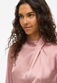 Blusa de seda rosa con un alto cuello y detalle fruncido en el frente. Presenta una textura suave y un caído ligero, adecuada para ocasiones formales.