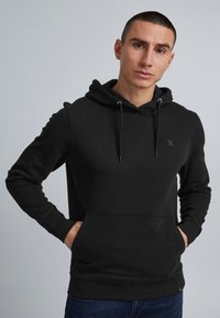 Man in een zwarte hoodie met een voorzak en trekkoorden, staand tegen een effen grijze achtergrond, handen in de zak.