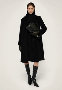 Černý kabát z vlněné směsi s propracovaným střihem, délka po kolena. Kombinován s černými koženými rukavicemi a špičatými kozačkami po kolena. Drží černou kabelku.