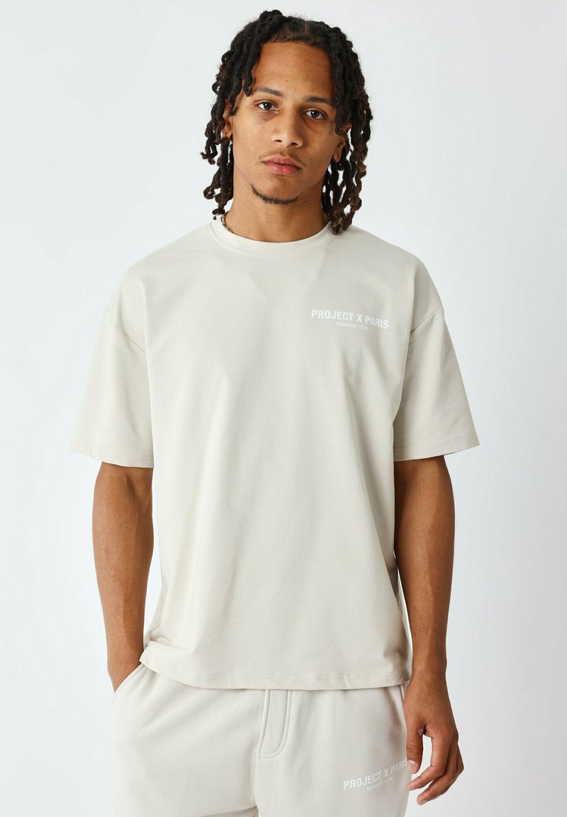 Camiseta de manga corta beige en un corte relajado con cuello redondo acanalado. Presenta "PROJECT X PARIS" impreso en blanco en el lado izquierdo del pecho.