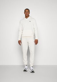 Sudadera blanca con un bolsillo tipo canguro y un pequeño logo en el pecho, combinada con pantalones jogger blancos a juego y zapatillas en blanco y negro.