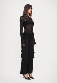 Top lungo a maniche in pizzo nero con righe texturizzate, sovrapposto a un bustier aderente; abbinato a pantaloni cargo neri e décolleté neri a punta.
