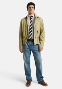 Lysbeige bomuldsjakke med lynlås og lommer, parret med enternet skjorte og stribet slips. Båret med lyseblå jeans og brune sko.