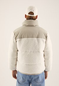Puffer jacket with white shearling texture, beige nylon shoulders, high collar, and relaxed fit. Paired with light blue jeans.