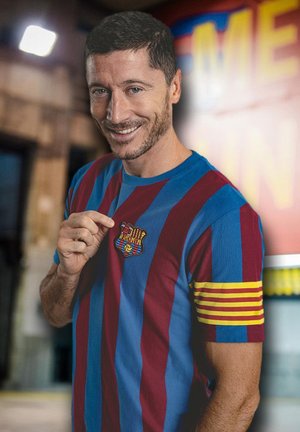Man lacht en wijst naar het FC Barcelona-badge op zijn blauw-rood gestreepte shirt met aanvoerdersbandje in een wazige binnenomgeving.