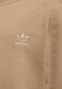 Béžová mikina Adidas s okrúhlym výstrihom, rebrovanou textúrou a vyšívaným logom na ľavej strane hrude.
