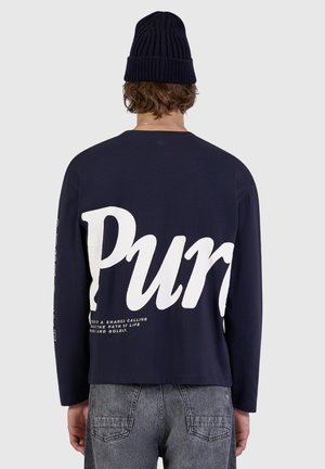 Person trägt ein marineblaues Langarmshirt mit großem weißem "Purs" Text auf dem Rücken, graue Jeans und eine dunkle Strickmütze, Rückansicht.