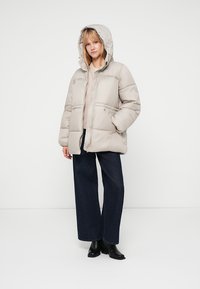 Chaqueta tipo puff en beige claro con capucha, con cremallera frontal y dos bolsillos frontales. Combinada con jeans de pierna ancha oscuros y botines negros.