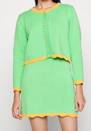 Kvinde iført et grønt strikket cardigan- og nederdelssæt med orange kant og knapper, stående mod en ensfarvet hvid baggrund.