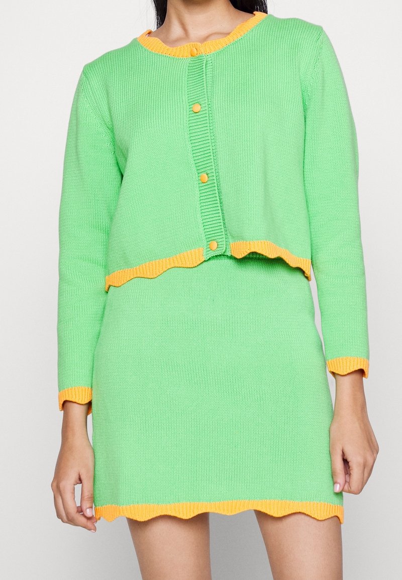 Femme portant un ensemble en tricot vert composé d'un cardigan et d'une jupe avec des bordures et des boutons orange, debout devant un fond blanc uni.