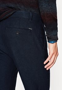 Granatowe, tkane spodnie o fakturowym wykończeniu, z tylną kieszenią z guzikem oraz małą metką marki. Dopasowana talia i gładki materiał.