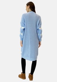 Cardigan lavorato a maglia blu chiaro con vestibilità morbida, caratterizzato da motivi a cuore bianchi sulle maniche. Polsini e orlo a costine. Indossato con leggings neri.